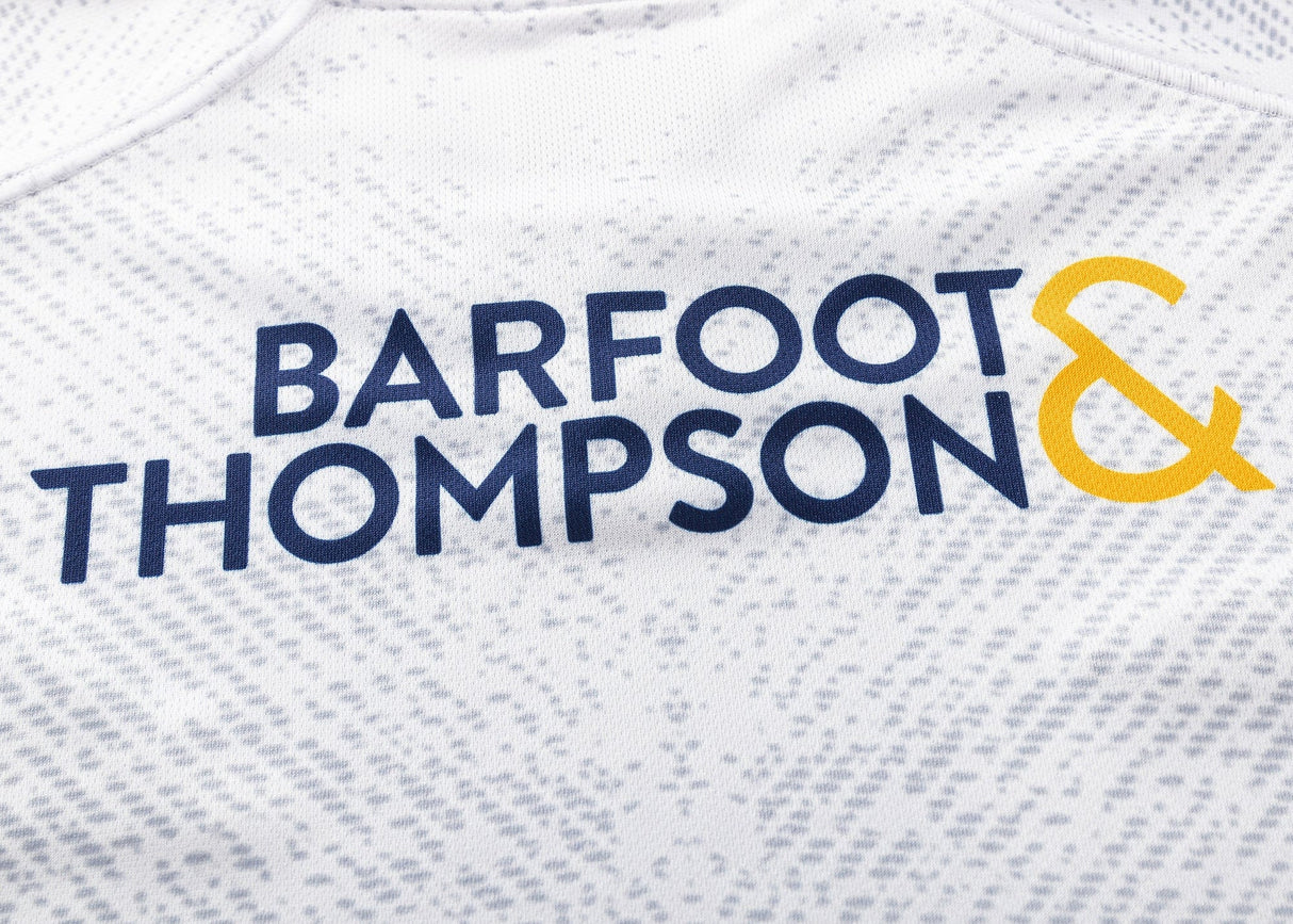 Close-up of a white fabric featuring "Barfoot & Thompson" in blue and yellow letters, echoing the stylish design of the Blues Super Rugby 25/26 Replica Away Jersey by Classic Sportswear.