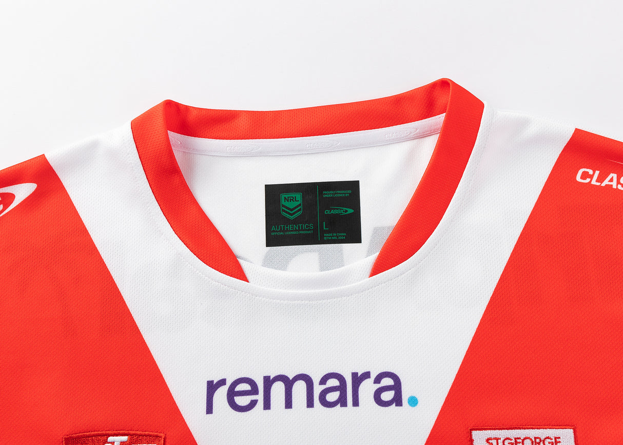 A Dragons NRL 25/26 Replica Home Jersey by Classic Sportswear is showcased. The jersey's red and white design includes a red collar with size and branding labels by Classic Sportswear inside, complete with sponsor logos for authenticity.