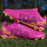 A pair of IDA Sports Helia Club FG women's soccer cleats in dark pink with gold accents and "IDA" printed in black, resting on green grass—these firm ground boots combine style and performance.