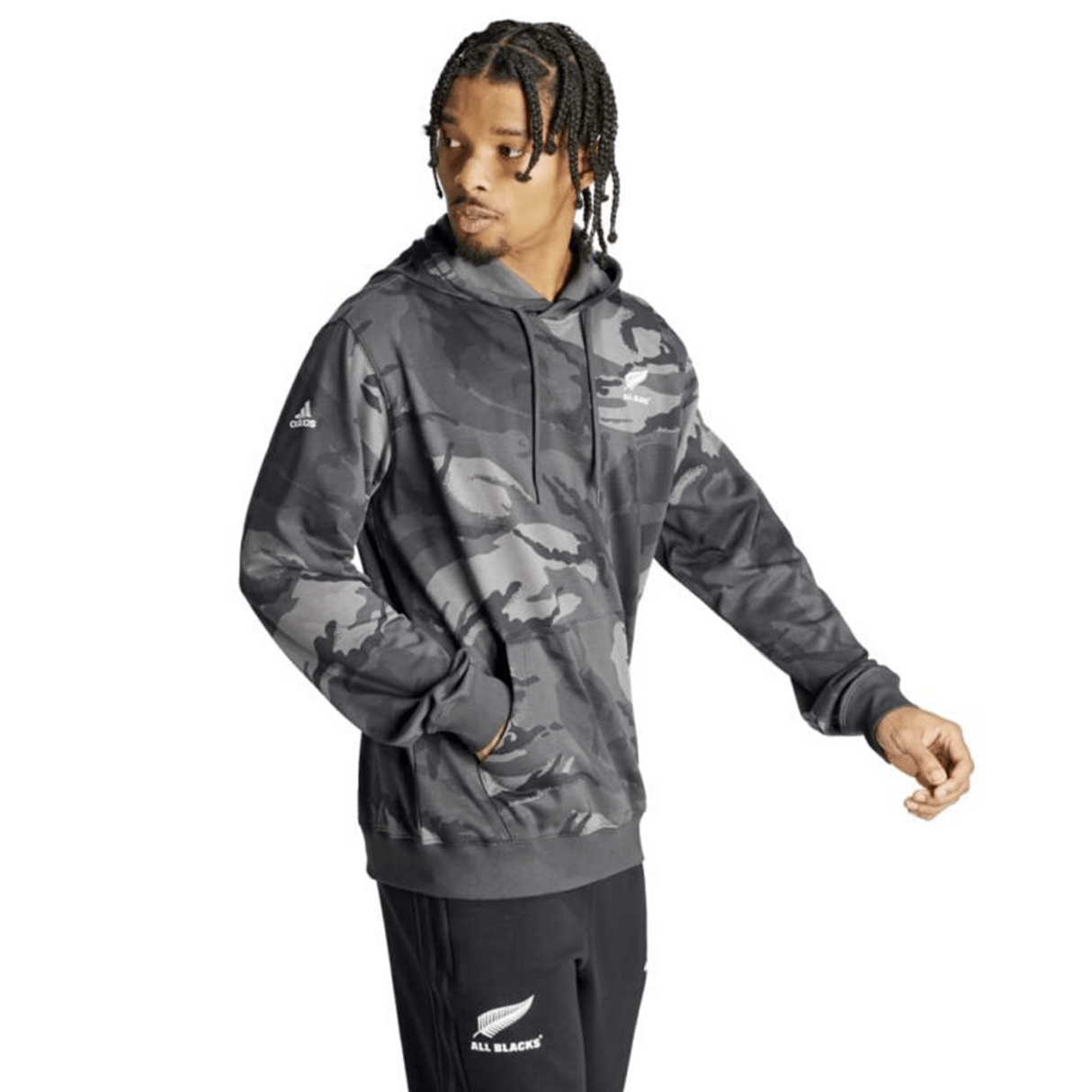 A person with braided hair is wearing an All Blacks Camo Hoodie by adidas, crafted from recycled polyester, paired with black track pants. The hoodie features a camouflage design along with the Adidas and All Blacks logos, and the individual is looking to the side.