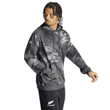 A person with braided hair is wearing an All Blacks Camo Hoodie by adidas, crafted from recycled polyester, paired with black track pants. The hoodie features a camouflage design along with the Adidas and All Blacks logos, and the individual is looking to the side.