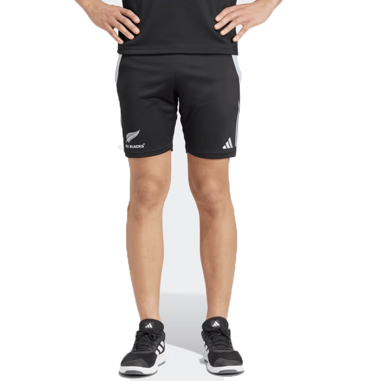 A person stands confidently, hands on hips, wearing sleek black "All Blacks 24/25 Performance Gym Shorts by Adidas," which feature logos reminiscent of All Blacks rugby shorts. The look is completed with a matching black t-shirt and sneakers, all crafted from recycled materials for a sustainable edge.
