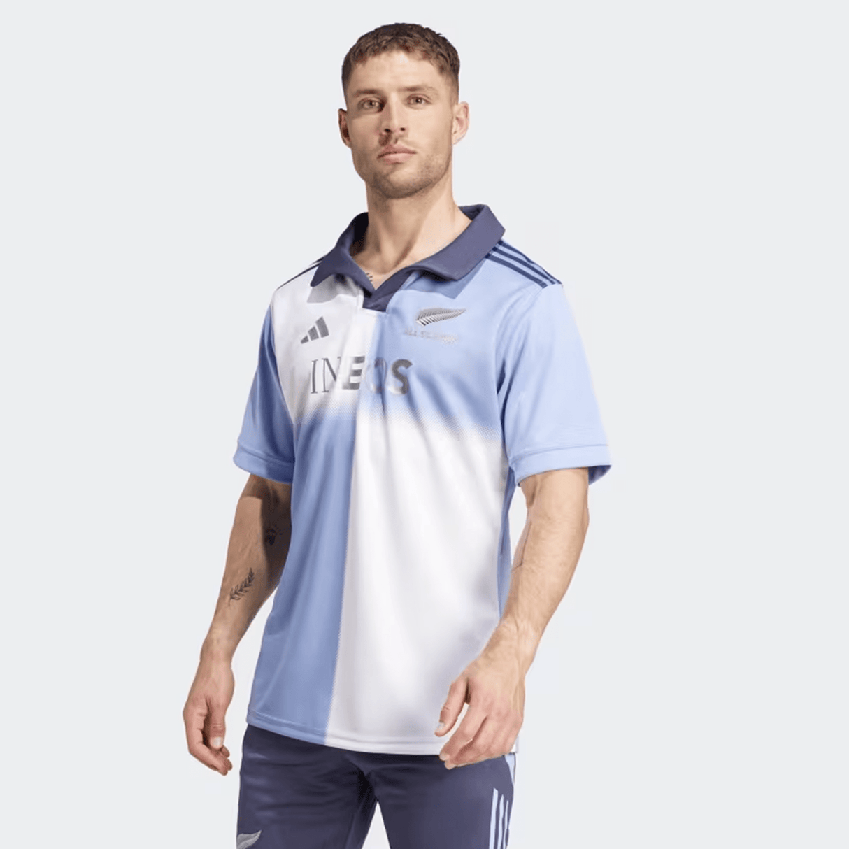 A man wearing the All Blacks 24/25 Training Jersey by adidas, featuring light blue and white colors and the silver fern logo, stands against a plain background with dark blue shorts.