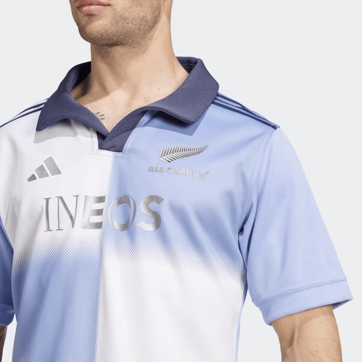 Someone wears the adidas All Blacks 24/25 Training Jersey in a light blue and white gradient with a striking collar, featuring the silver fern and logos of adidas and Ineos on the front.