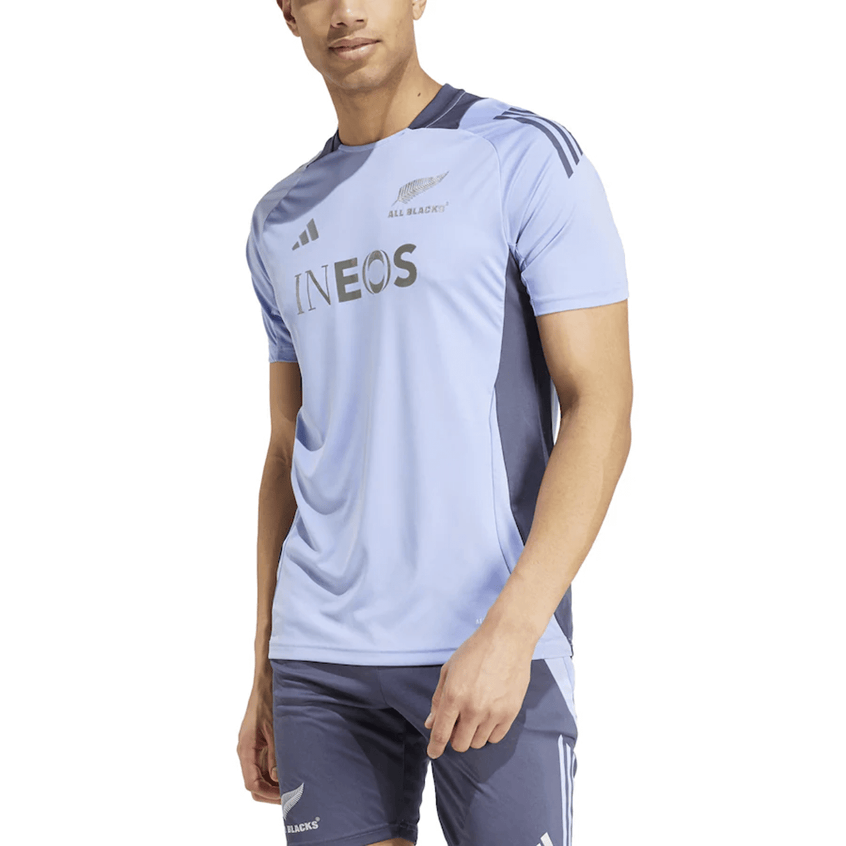 A person is wearing a light blue and dark gray sports jersey and shorts featuring the word "INEOS" and three stripes on the sleeves, similar to the All Blacks 24/25 Performance T-Shirt by adidas.