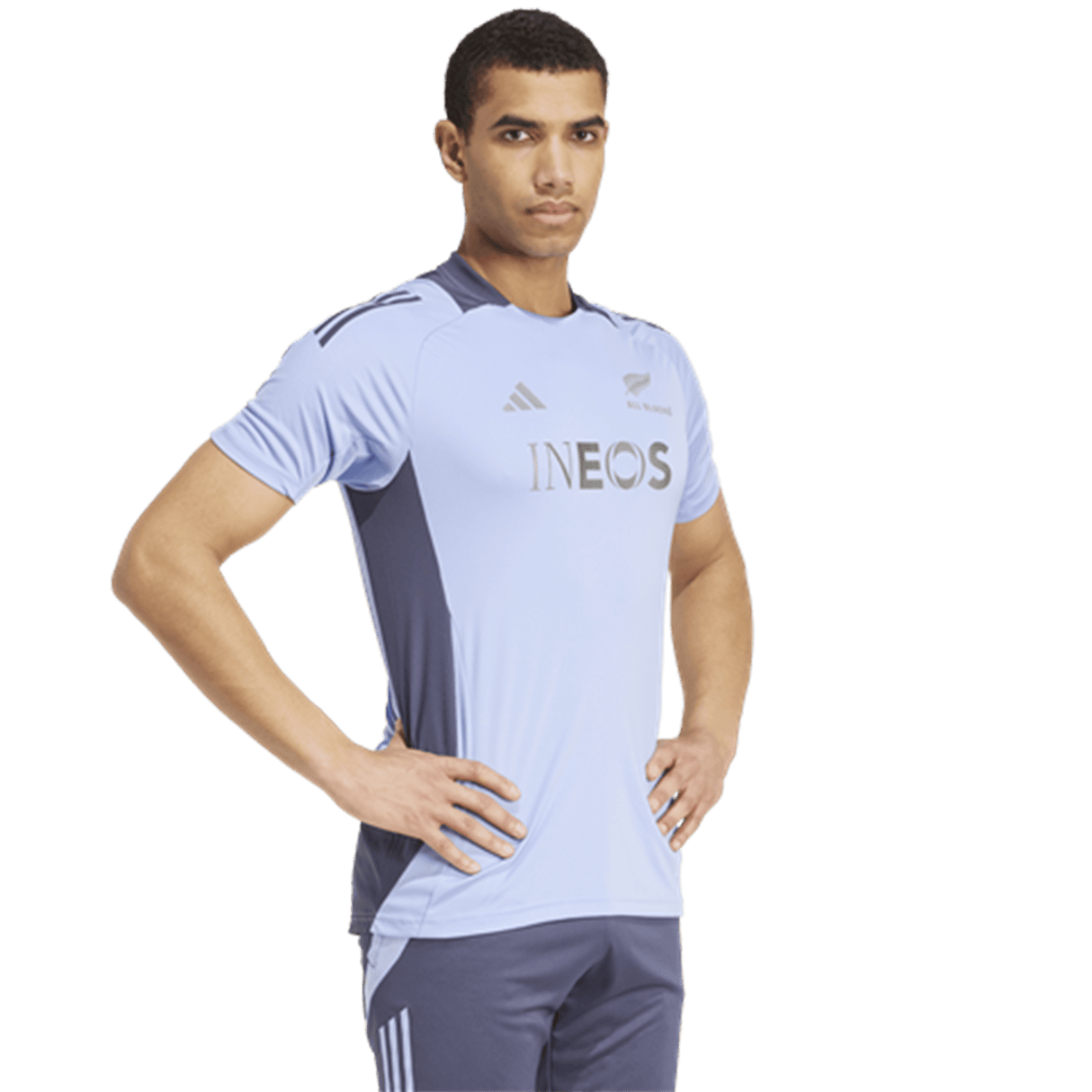 A person wearing the All Blacks 24/25 Performance T-Shirt by adidas, featuring a light blue and dark blue design with an "INEOS" logo, stands with hands on hips and looks forward; the AEROREADY technology in the fabric helps them stay cool and dry, capturing the resilience of the All Blacks.