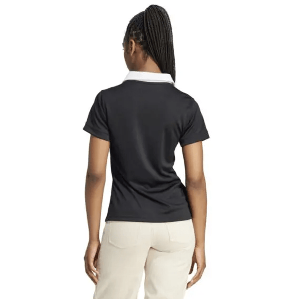 A woman with braided hair stands with her back to the camera, wearing an adidas Women's All Blacks 24/25 Home Supporters Jersey featuring AEROREADY technology, paired with beige pants.