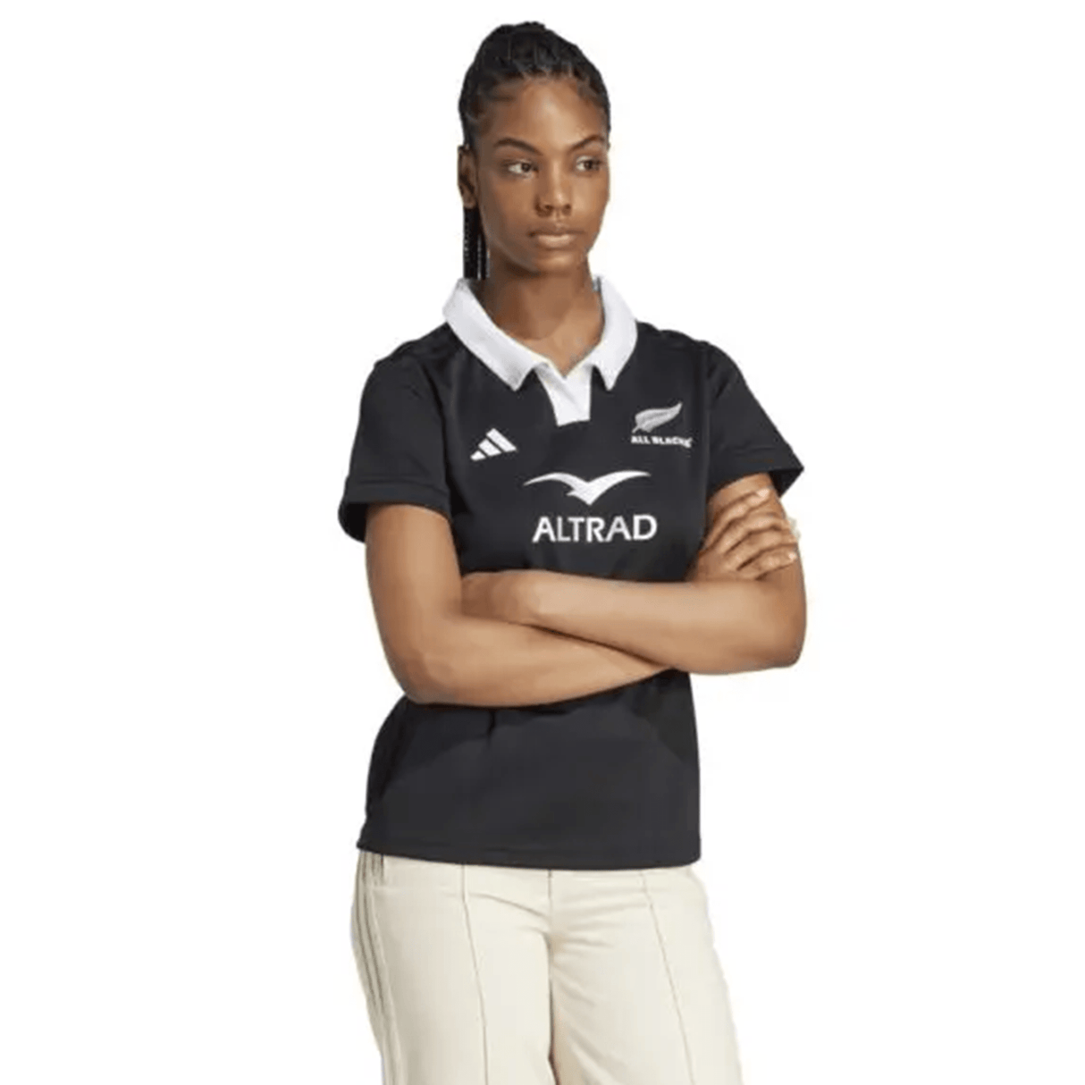 With arms crossed, the individual wears an adidas Women's All Blacks 24/25 Home Supporters Jersey made with AEROREADY technology, paired with light-colored pants against a plain background.