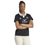 With arms crossed, the individual wears an adidas Women's All Blacks 24/25 Home Supporters Jersey made with AEROREADY technology, paired with light-colored pants against a plain background.