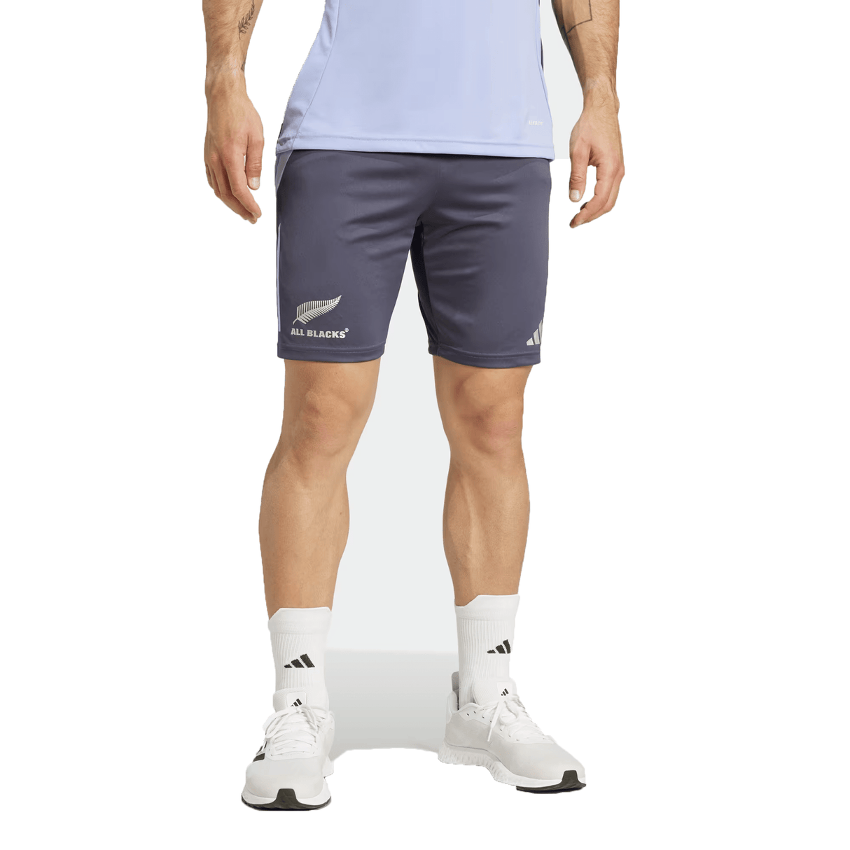A man dressed in a light blue sports shirt and the dark All Blacks 24/25 Performance Gym Shorts by adidas, made from recycled materials and featuring the "All Blacks" logo, stands against a plain background, complemented by white sneakers and socks.