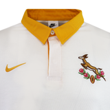The Springboks Heritage Long Sleeve Rugby Jersey by Nike features a white base, yellow collar, yellow Nike logo, and embroidered Springbok with flowers—a stylish tribute to classic rugby jersey designs.