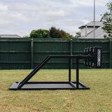An IronBack Ironback Gorilla 8 Man Scrum Machine in black metal stands on a grassy lawn, with a green wooden fence and houses in the background.