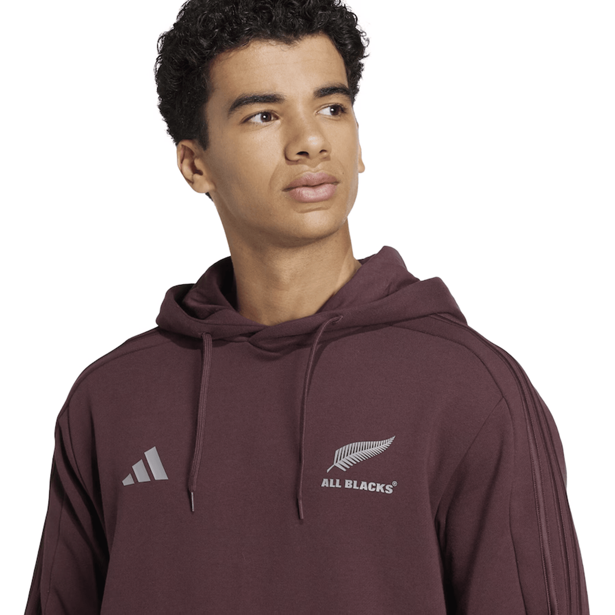 Wearing the adidas All Blacks 24/25 Fan Rugby Hoodie, featuring the iconic adidas and All Blacks logos in a striking maroon, this individual looks slightly to the side against a plain background—a must-have addition for any New Zealand rugby fan’s apparel collection.