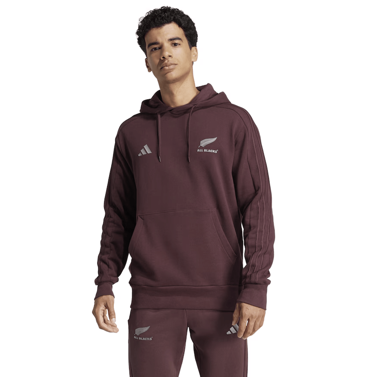 A man stands facing forward against a plain white background, wearing the maroon All Blacks 24/25 Fan Rugby Hoodie by adidas along with matching joggers featuring adidas logos. This stylish adidas hoodie is an ideal addition to any New Zealand rugby fan’s apparel collection.