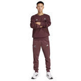 An individual in an adidas All Blacks 24/25 Fan Crewneck Sweatshirt and sleek white sneakers strikes a confident pose against a light gray backdrop.