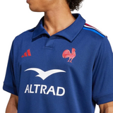 Someone wears the adidas France FFR 25/26 Replica Home Jersey, featuring a blue design, colored shoulder stripes, a red rooster emblem, and "ALTRAD" plus another white logo on the front.