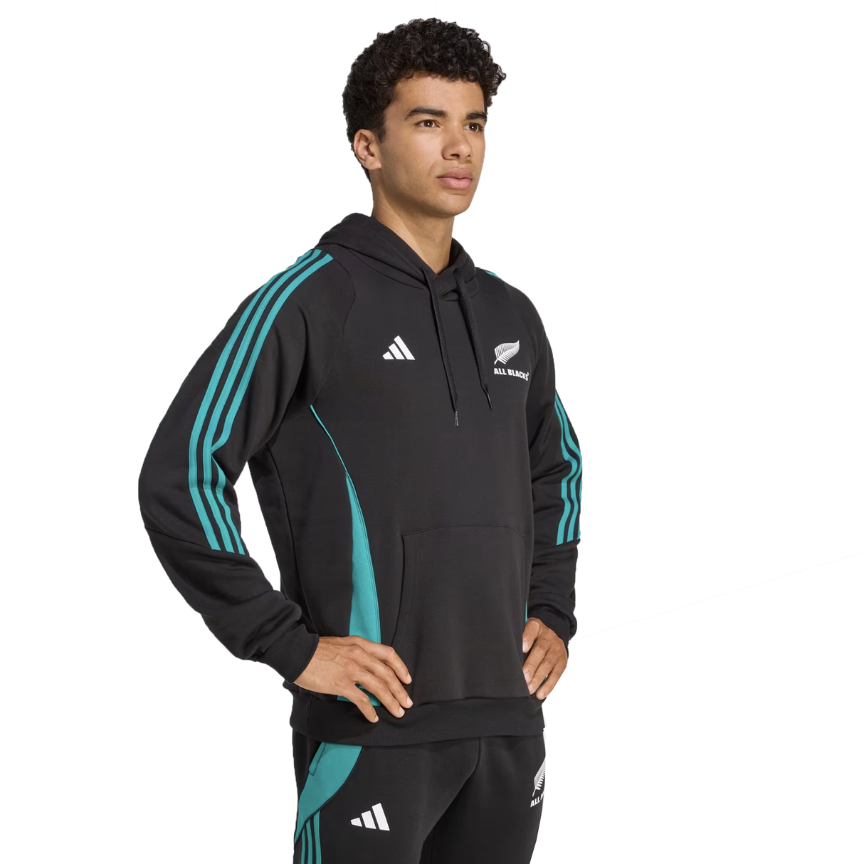 A person stands with hands on hips, wearing the black and teal All Blacks 25/26 Training Hoodie by adidas and matching pants, both featuring New Zealand rugby logos.