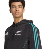 A young man wears the All Blacks 25/26 Training Hoodie by adidas, featuring teal stripes and the All Blacks rugby team logo on the chest.