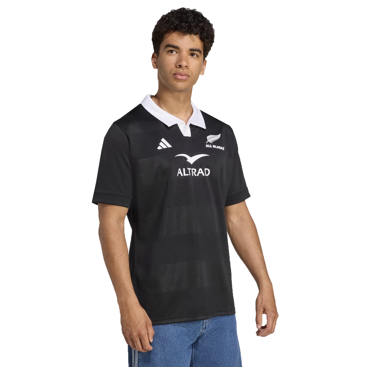 A person models the adidas All Blacks 25/26 Replica Home Jersey in black with a white collar and logos, made from recycled polyester, paired with blue jeans, standing slightly sideways against a plain background.