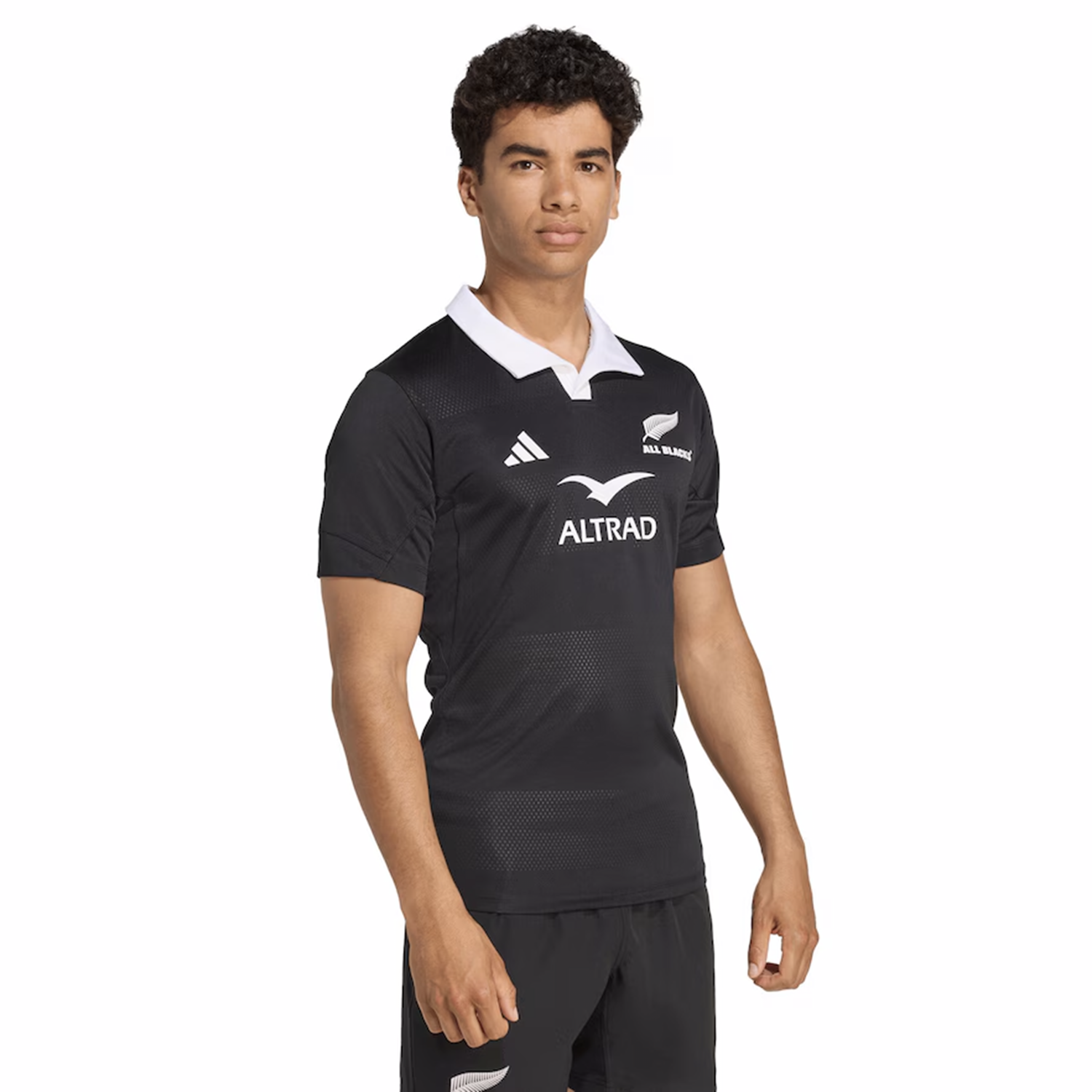 A person wears the All Blacks 25/26 Performance Home Jersey by adidas, a black rugby jersey with a white collar and sponsor logos, standing against a plain white background.