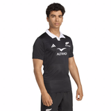 A person wears the All Blacks 25/26 Performance Home Jersey by adidas, a black rugby jersey with a white collar and sponsor logos, standing against a plain white background.