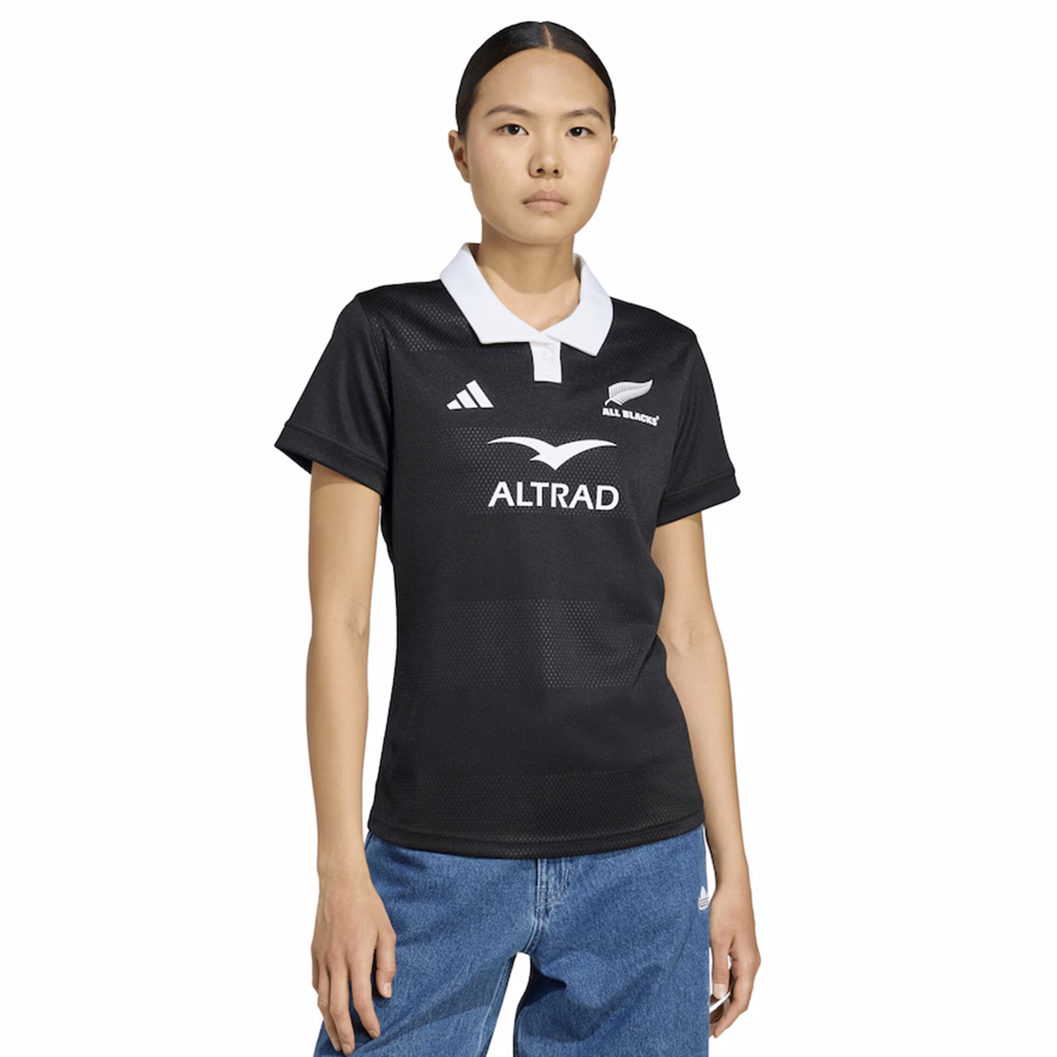 The Women's All Blacks 25/26 Replica Home Jersey by adidas features a black design with a white collar and logo, styled here with blue jeans for a classic rugby look against a simple background.
