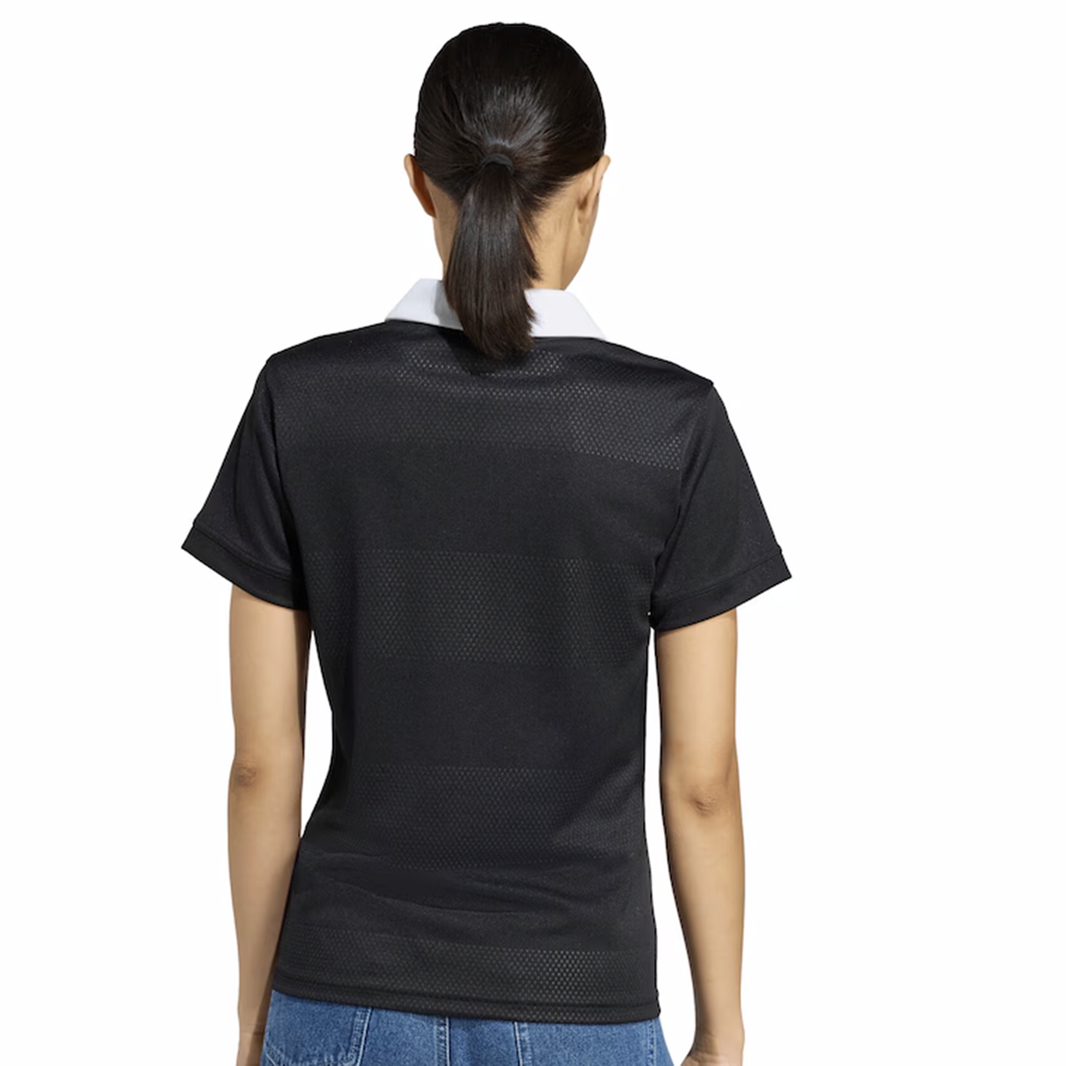 A person with dark hair in a ponytail wears the adidas Women's All Blacks 25/26 Replica Home Jersey and blue jeans, facing away from the camera against a white background.
