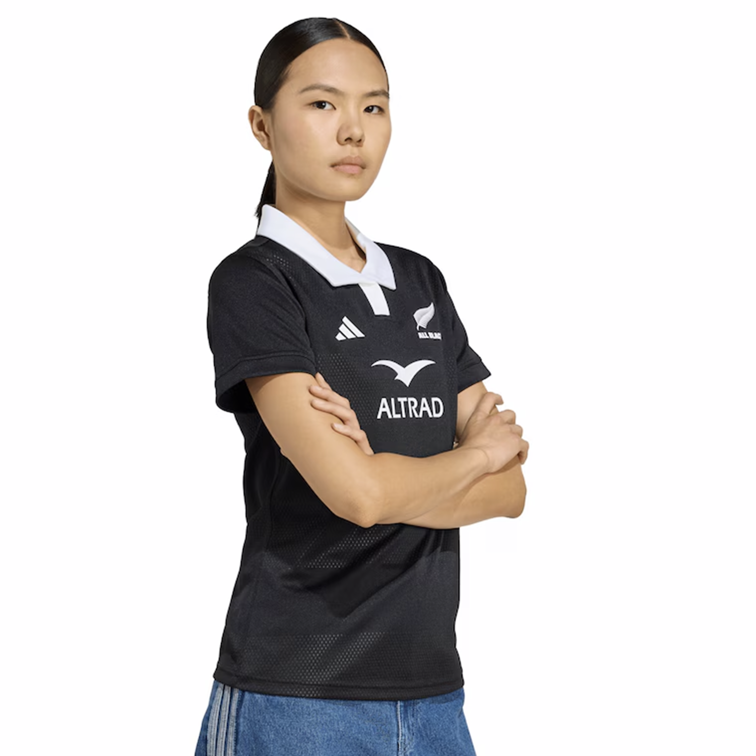 A person stands with arms crossed, wearing the adidas Women's All Blacks 25/26 Replica Home Jersey with a white collar and logos, paired with blue jeans, facing slightly to the side against a white background.