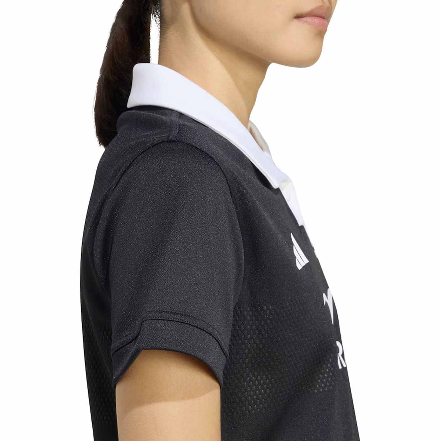 A woman is shown from the shoulders up in side profile, wearing the adidas Women's All Blacks 25/26 Replica Home Jersey, a black collared sports shirt with white trim.