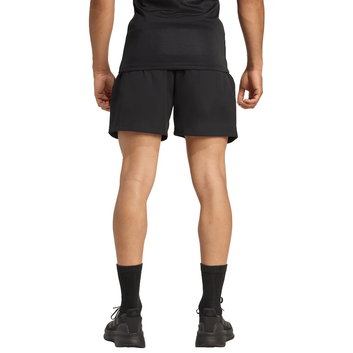 A person is standing facing away from the camera, wearing a black shirt, adidas All Blacks 25/26 Replica Home Shorts, black socks, and black shoes.
