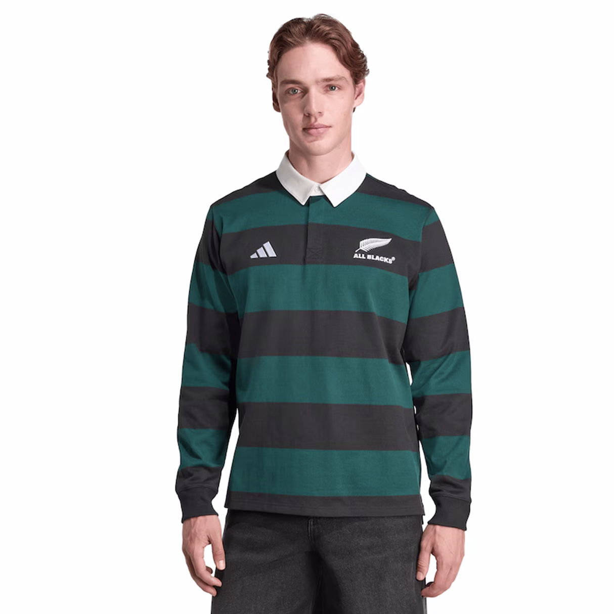 A young man stands facing forward, wearing the adidas All Blacks 25/26 Long Sleeve Heritage Polo, featuring green and black stripes, a white collar, and the New Zealand All Blacks logo.