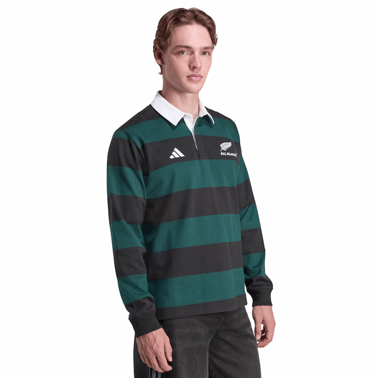 A person wears the All Blacks 25/26 Long Sleeve Heritage Polo by adidas, featuring green and black stripes, a white collar, and New Zealand and adidas logos, standing against a plain background.