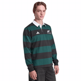 A person wears the All Blacks 25/26 Long Sleeve Heritage Polo by adidas, featuring green and black stripes, a white collar, and New Zealand and adidas logos, standing against a plain background.