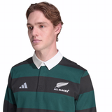 Someone wearing the adidas All Blacks 25/26 Long Sleeve Heritage Polo, featuring green and black stripes, a white collar, the adidas logo, and the "ALL BLACKS" insignia.
