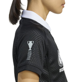 A person wears the Women's Black Ferns WRWC25 Replica Home Jersey by adidas, a black collared sports shirt with a white collar and sleeve trophy graphic listing years, inspired by the official Black Ferns kit.