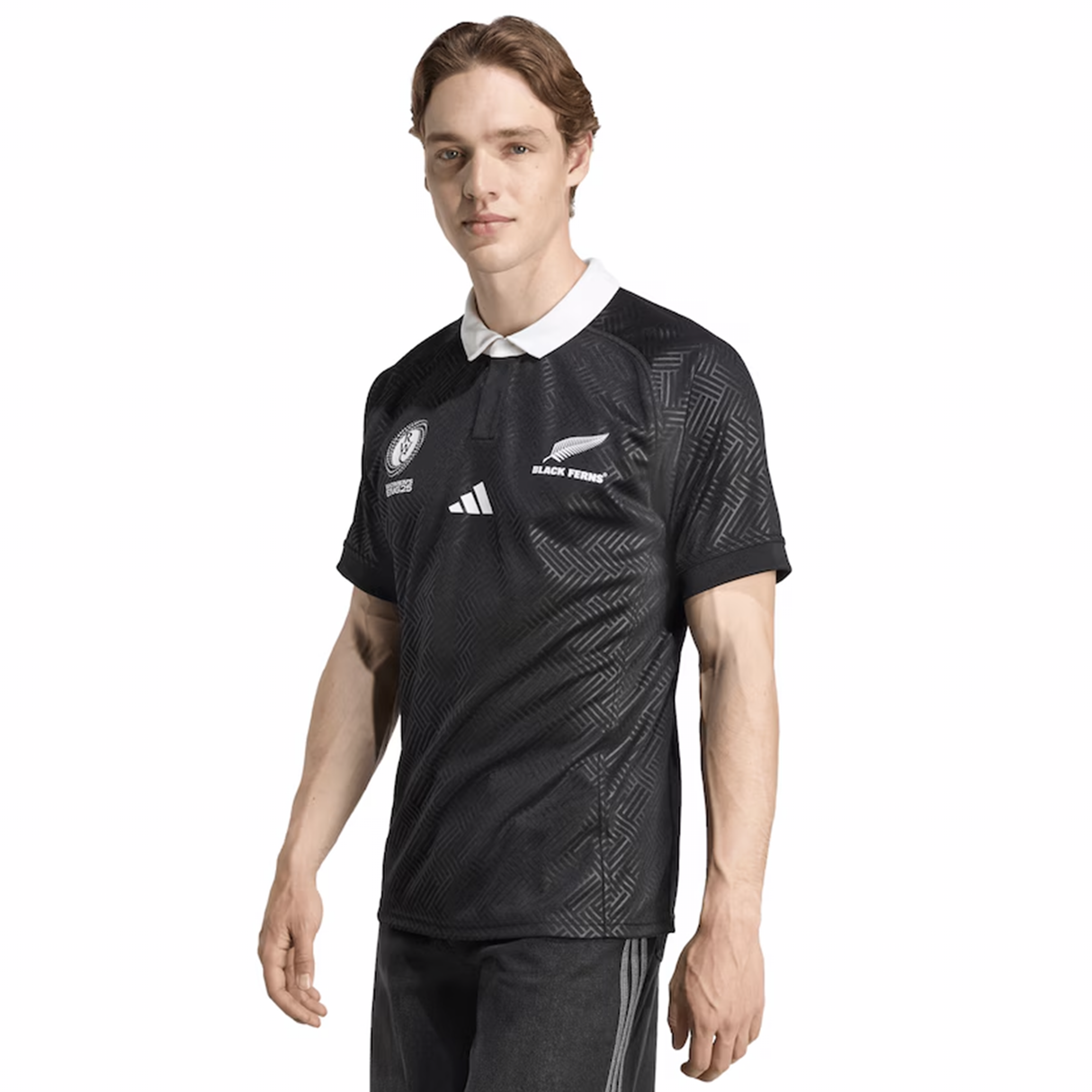 A man stands facing forward with one arm relaxed by his side, wearing the adidas Black Ferns WRWC25 Replica Home Jersey—a black collared rugby top featuring white logos and a geometric pattern.