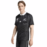 A man stands facing forward with one arm relaxed by his side, wearing the adidas Black Ferns WRWC25 Replica Home Jersey—a black collared rugby top featuring white logos and a geometric pattern.