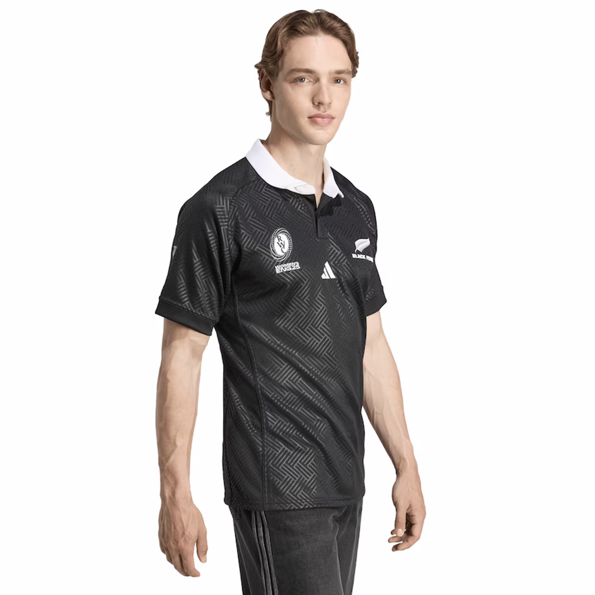 A man models the adidas Black Ferns WRWC25 Replica Home Jersey, featuring a black design with a white collar and branded logos, standing slightly to the side against a plain white background.