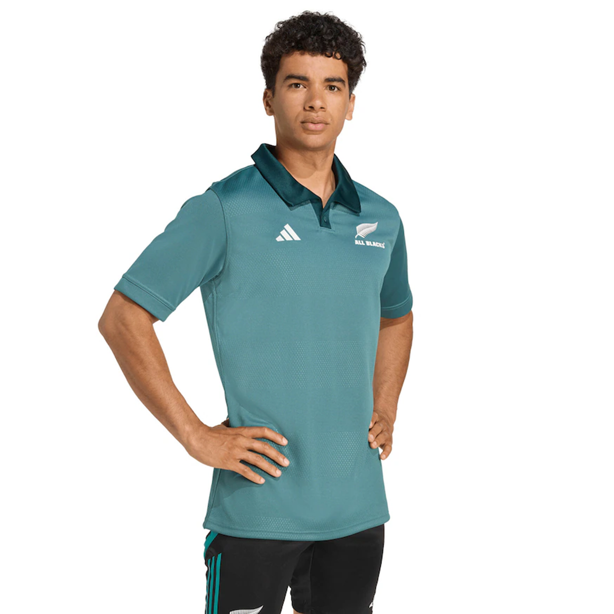 A person stands with hands on hips, wearing the teal All Blacks 25/26 Replica Training Jersey by adidas, which features sports logos, along with black pants, facing forward against a plain background.