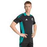 A young man stands with hands on hips, wearing the adidas All Blacks 25/26 Performance T-Shirt in black and teal with "New Zealand" logos, set against a plain white background.