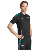 A person wears the adidas All Blacks 25/26 Training Polo with matching pants featuring teal accents, hands on hips against a white backdrop. This modern adidas polo highlights iconic All Blacks pride.