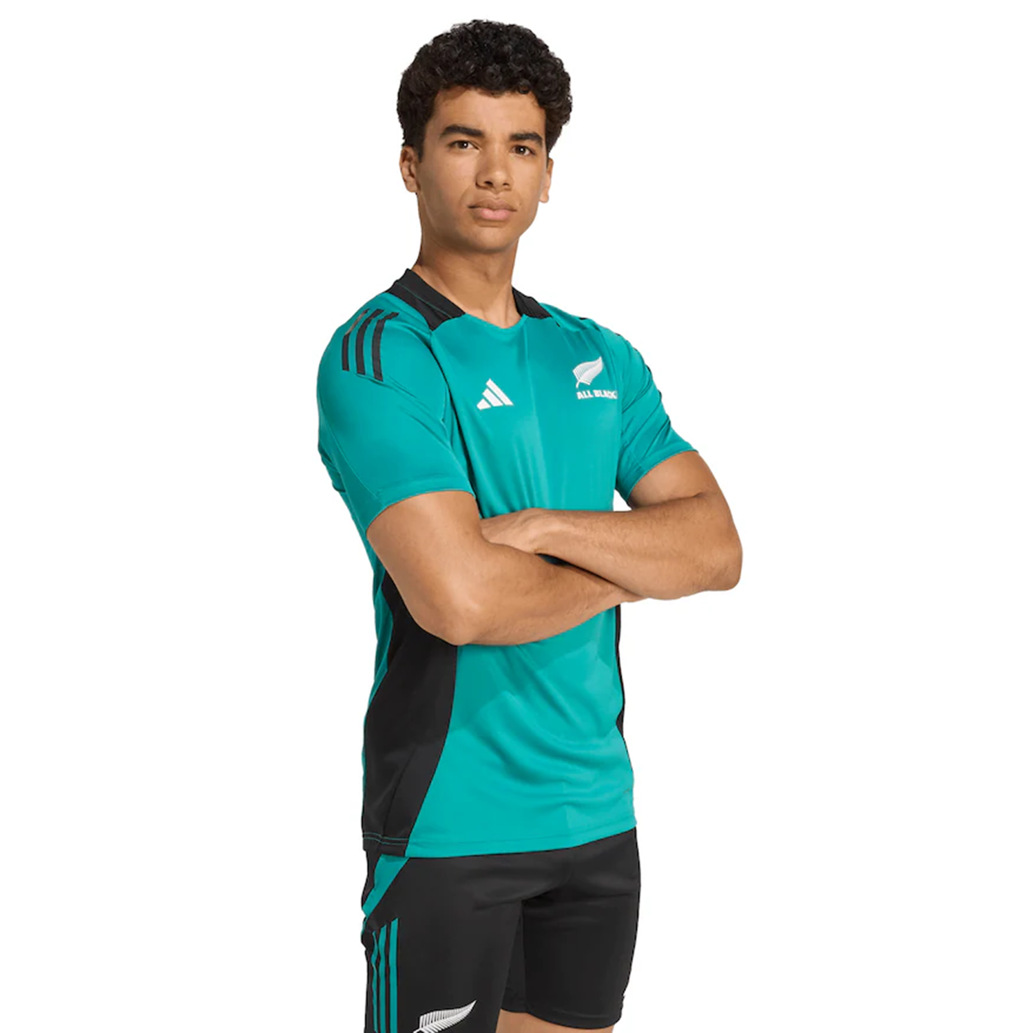 A person stands with arms crossed, wearing the All Blacks 25/26 Performance T-Shirt by adidas in teal and black, featuring All Blacks and adidas logos on the shirt and shorts.