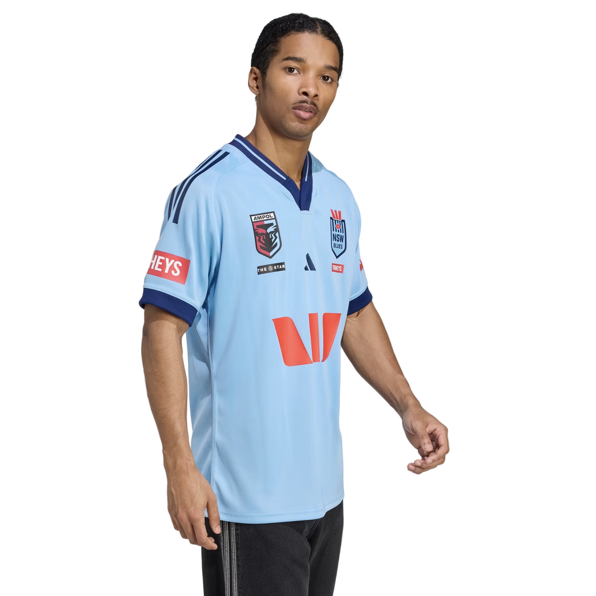 A man in the New South Wales Blues 25/26 Replica Home Jersey by adidas, featuring sponsor logos, stands against a plain background, facing slightly to the side.