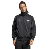 A person wears the adidas Originals Limited Edition All Blacks Firebird Track Top, featuring a black jacket with a silver fern logo, made from recycled polyester, set against a plain white background.