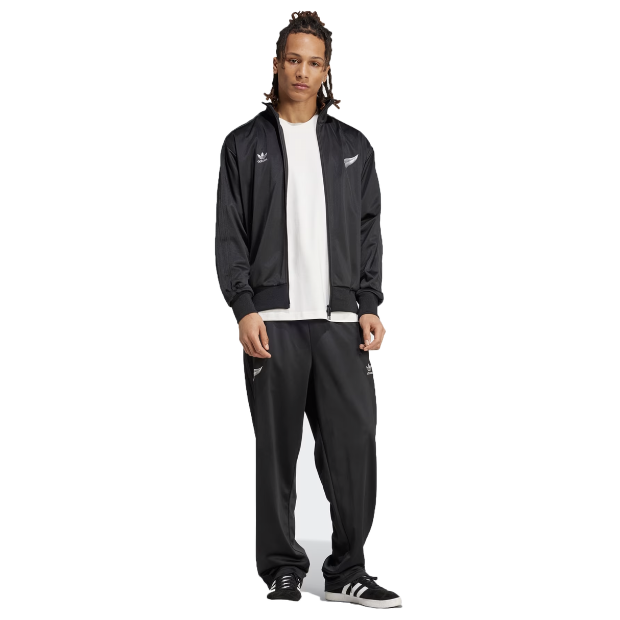 A person stands wearing the Originals Limited Edition All Blacks Firebird Track Top by adidas, crafted from recycled polyester, paired with a white shirt, black track pants, and black sneakers. The iconic adidas logo graces the look, merging style with sustainability.