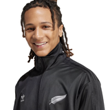 The person is smiling against a white background, wearing the Originals Limited Edition All Blacks Firebird Track Top by adidas, featuring the iconic adidas logo.