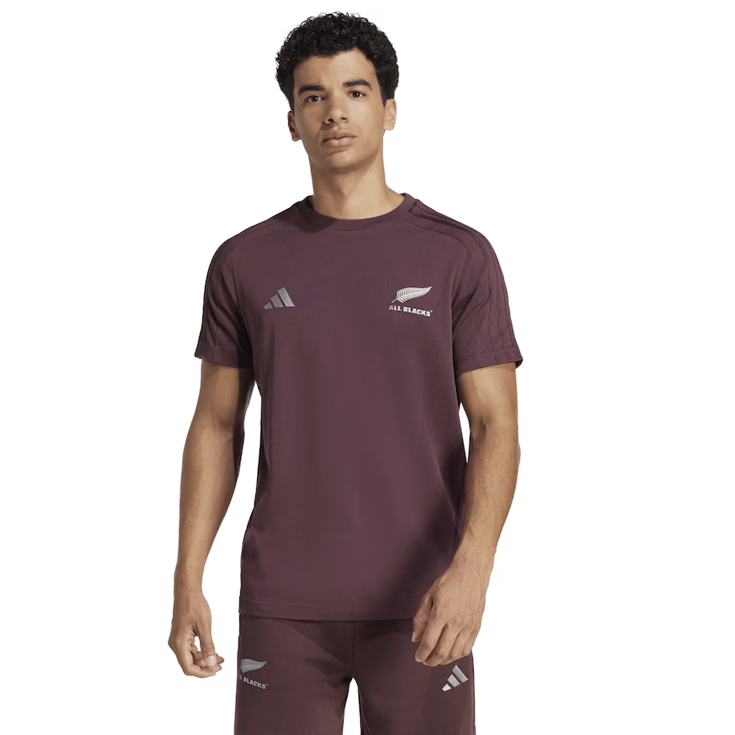 A person confidently poses against a white background wearing the maroon All Blacks 24/25 Fan Tee by adidas, featuring subtle 3-Stripes detailing and the "All Blacks" logo.