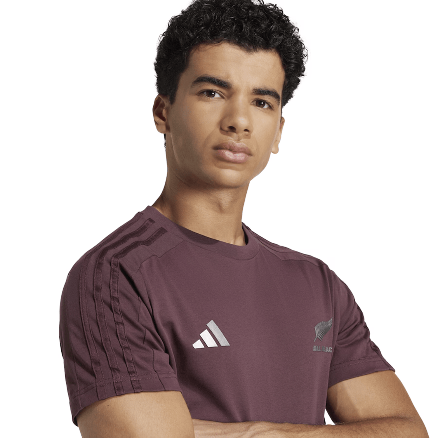 A person with curly hair crosses their arms and looks at the camera against a white background, wearing the All Blacks 24/25 Fan Tee by adidas.