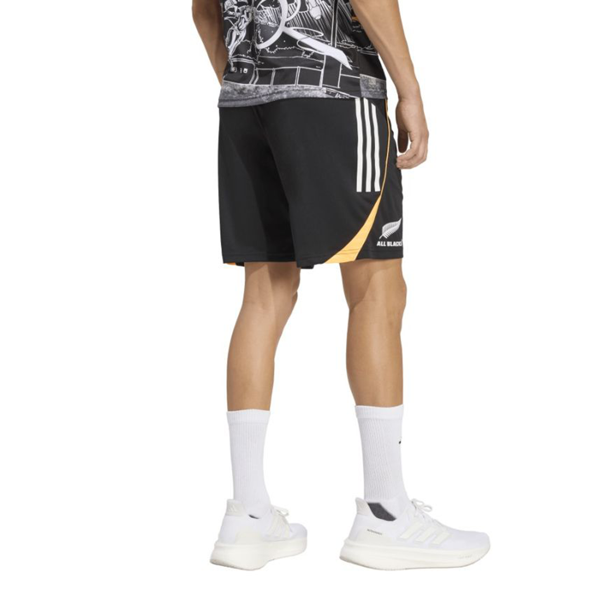 A person stands facing away, wearing adidas All Blacks 26/27 Marvel Performance Gym Shorts with white stripes, a graphic shirt, white crew socks, and white sneakers.