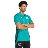 A man stands with arms crossed, wearing the adidas All Blacks 25/26 Training Polo in teal and black shorts with logos, facing slightly left against a plain background.